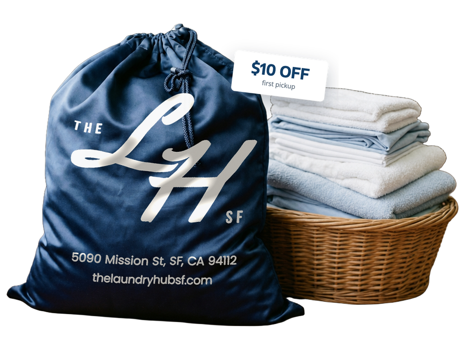 The Laundry Hub SF navy drawstring laundry bag, printed with the LH monogram and 5090 Mission St, beside a basket of freshly folded towels